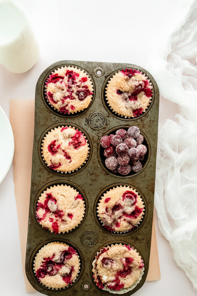Overhead shot of cranberry orange muffins baked in a vintage muffin tin, with sugared cranberries filling one of the cups. The muffins are golden and studded with vibrant red cranberries, set on a white marble-style background with soft fabric and a milk bottle in the frame.