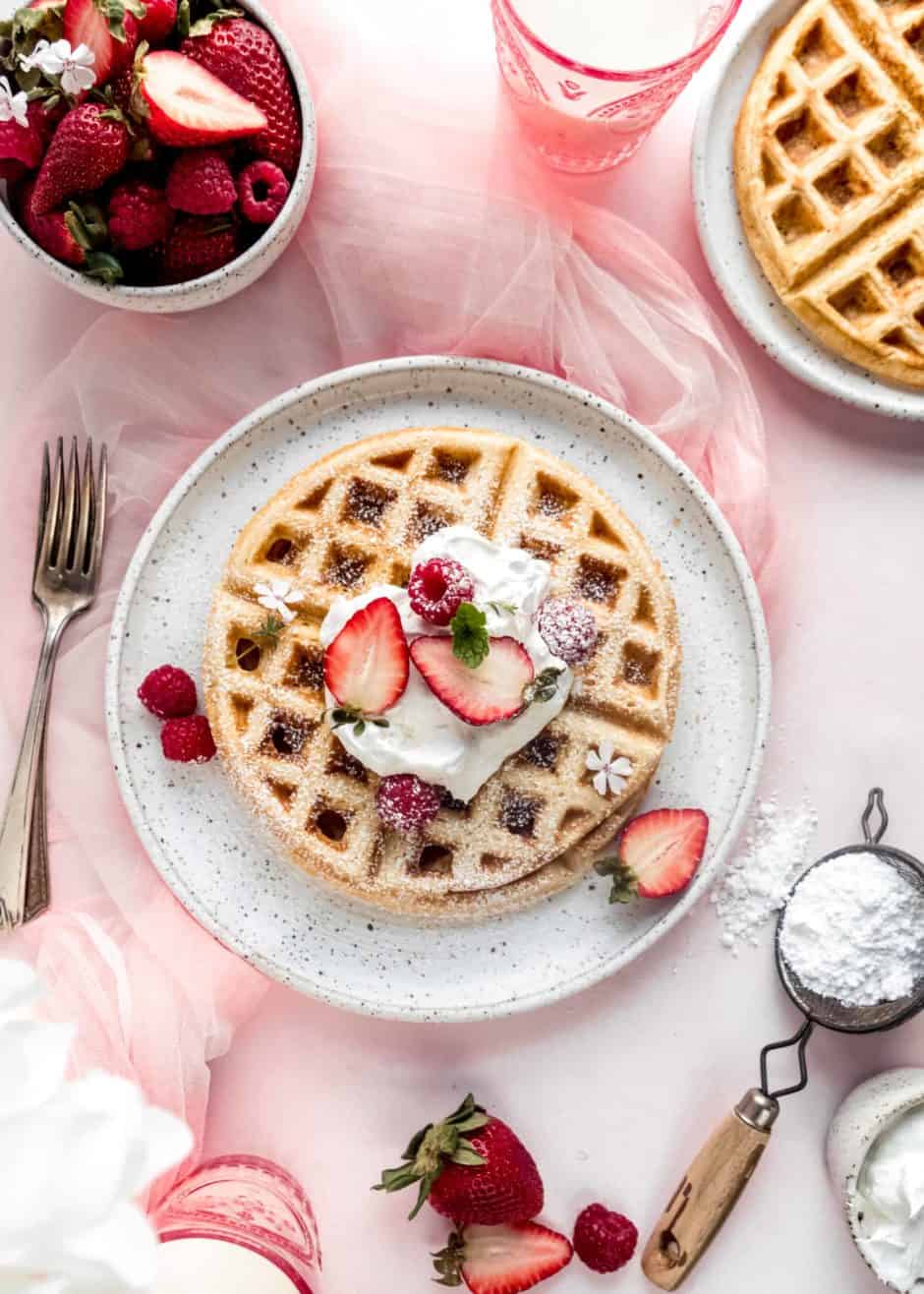 Fulffy yeast Belgian waffle on white plate topped with whipped cream and strawberries. The plate is sitting on a light pink background and surrounded by a bowl of strawberries, powdered sugar in a sifter, and a pink glass of milk