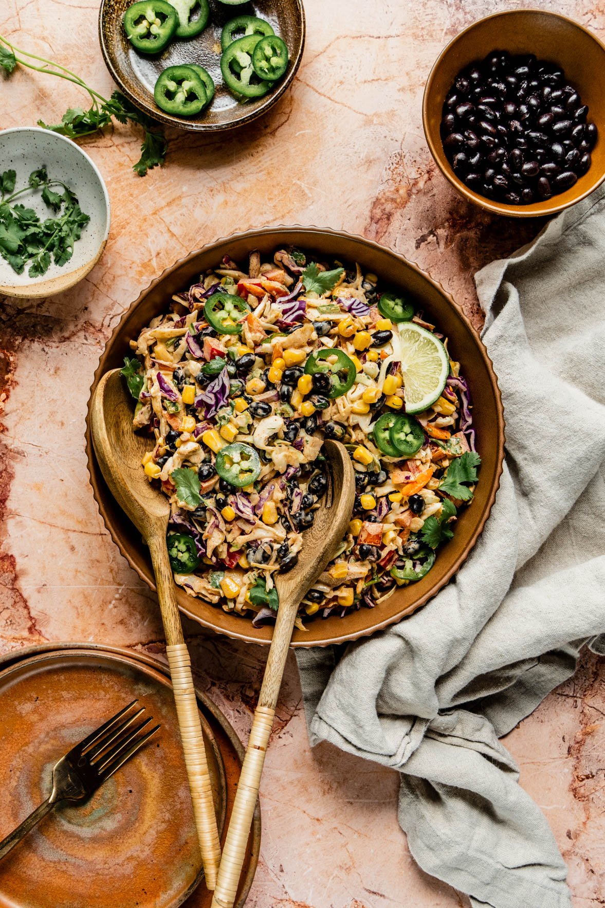 summer slaw in brown bowl on brown marble background with salad tongs in bowl.  Also on the counter is a bowl of black beans, a dish with sliced jalapeno and a bowl of fresh cilanto.