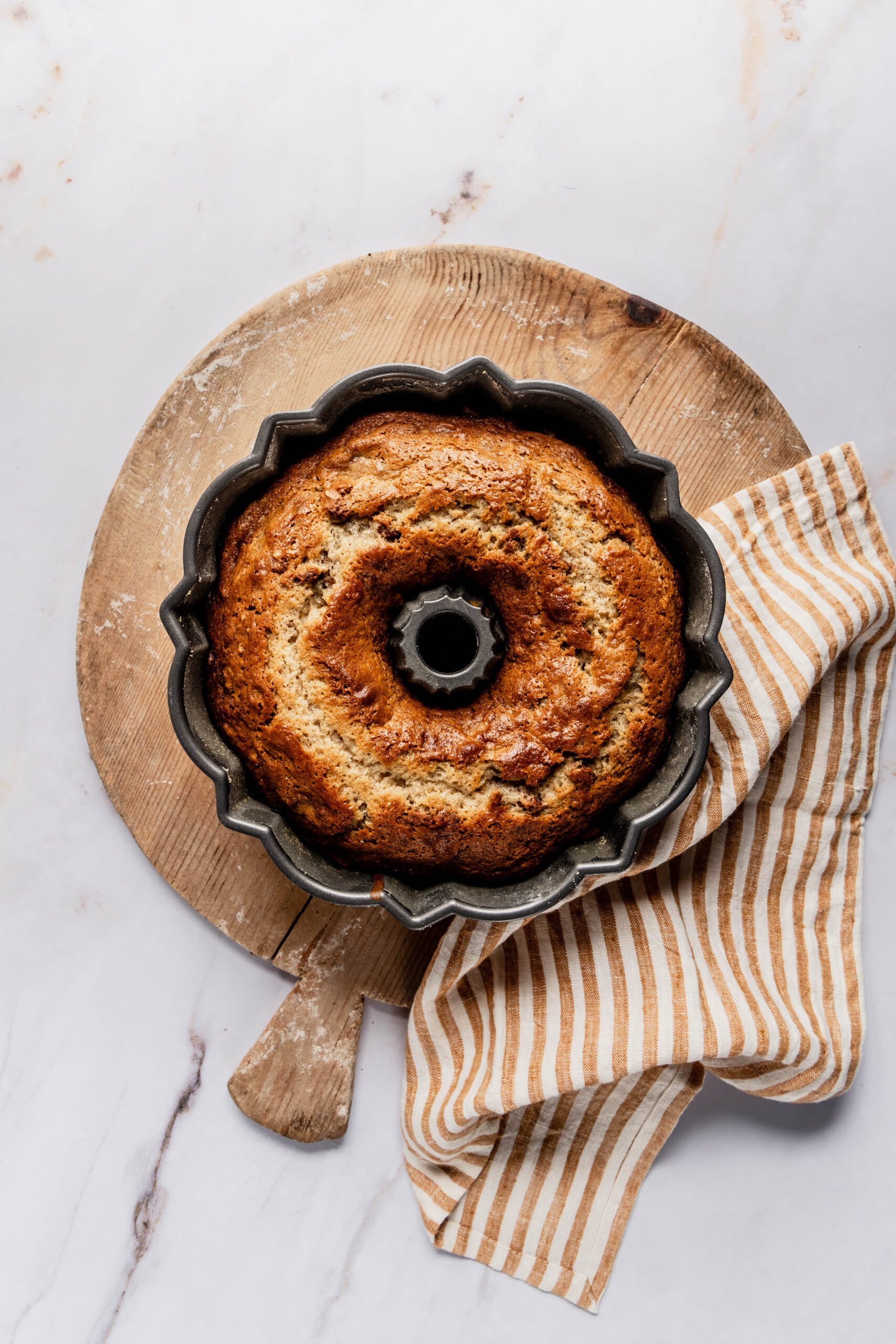 Banana nut bread bundt cake baked in bundt pan on round cutting board.  There's a yellow and white striped towel next to the bundt pan
