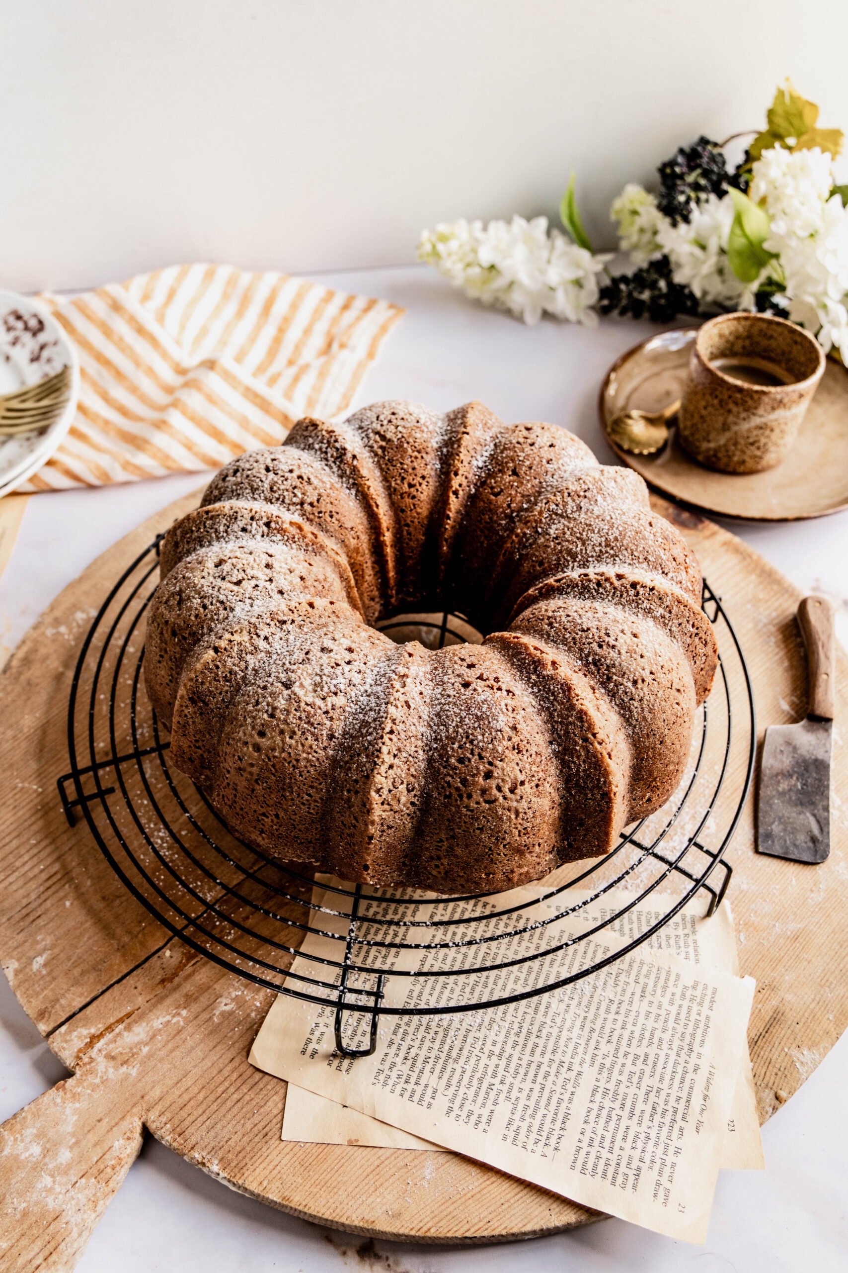 side shot of Banana nut bread bundt cake baked and removed from bundt pan on round cooling rack.  The cooling rack is sitting on top of the round cutting board and there's a cup of coffee with a gold spoon in the background. 