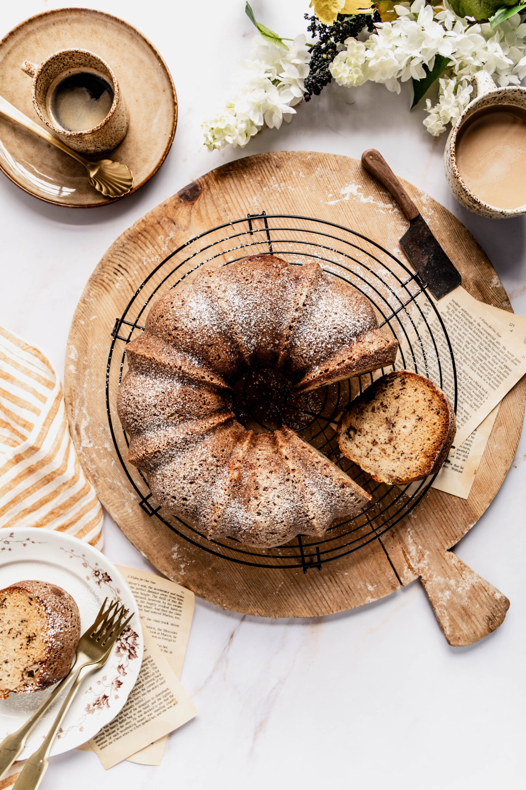 Banana nut bread bundt cake baked removed from bundt pan on cooling round cutting board with a slice taken out and placed on a plate. there's two cups of coffee above the cutting board and white flowers on the counter.