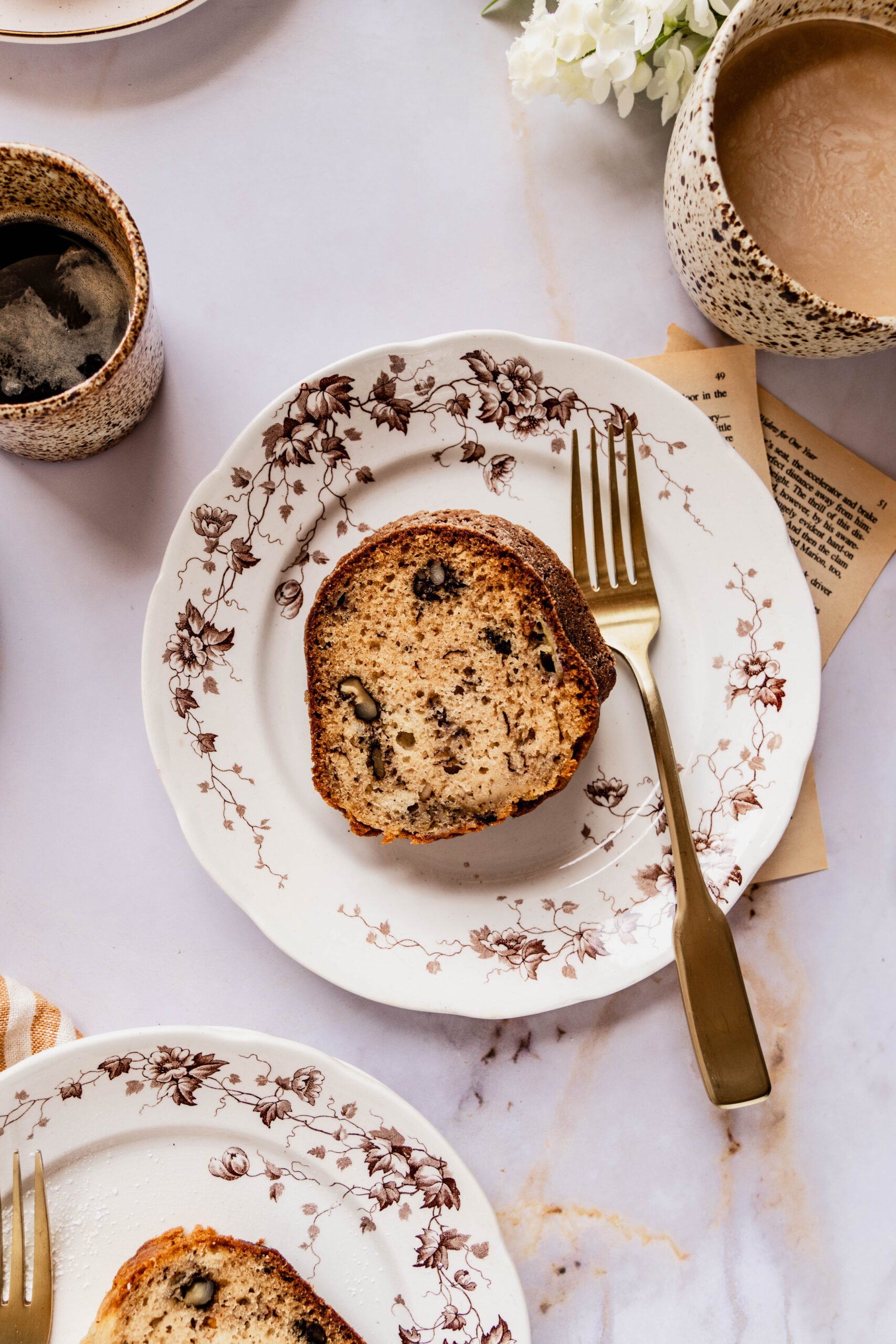 a close up shot of a sliced piece of Banana nut bread bundt cake on a white plate with two cups of coffee and white flowers on the marble counter.