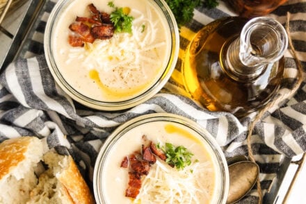 Overhead view of two jars filled with creamy Crockpot cauliflower soup, topped with shredded cheese, crispy bacon bits, fresh herbs, and a drizzle of olive oil, styled with crusty bread and spoons on a neutral cloth backdrop.