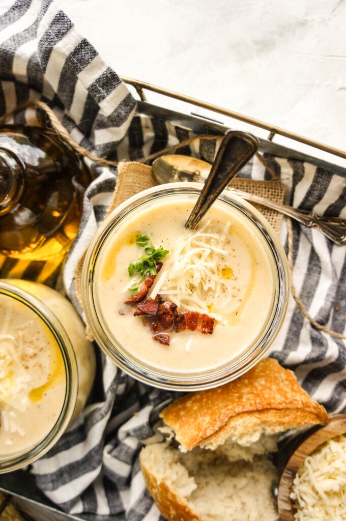 Overhead view of one jar filled with creamy Crockpot cauliflower soup, topped with shredded cheese, crispy bacon bits, fresh herbs, and a drizzle of olive oil, styled with crusty bread and spoons on a neutral cloth backdrop.