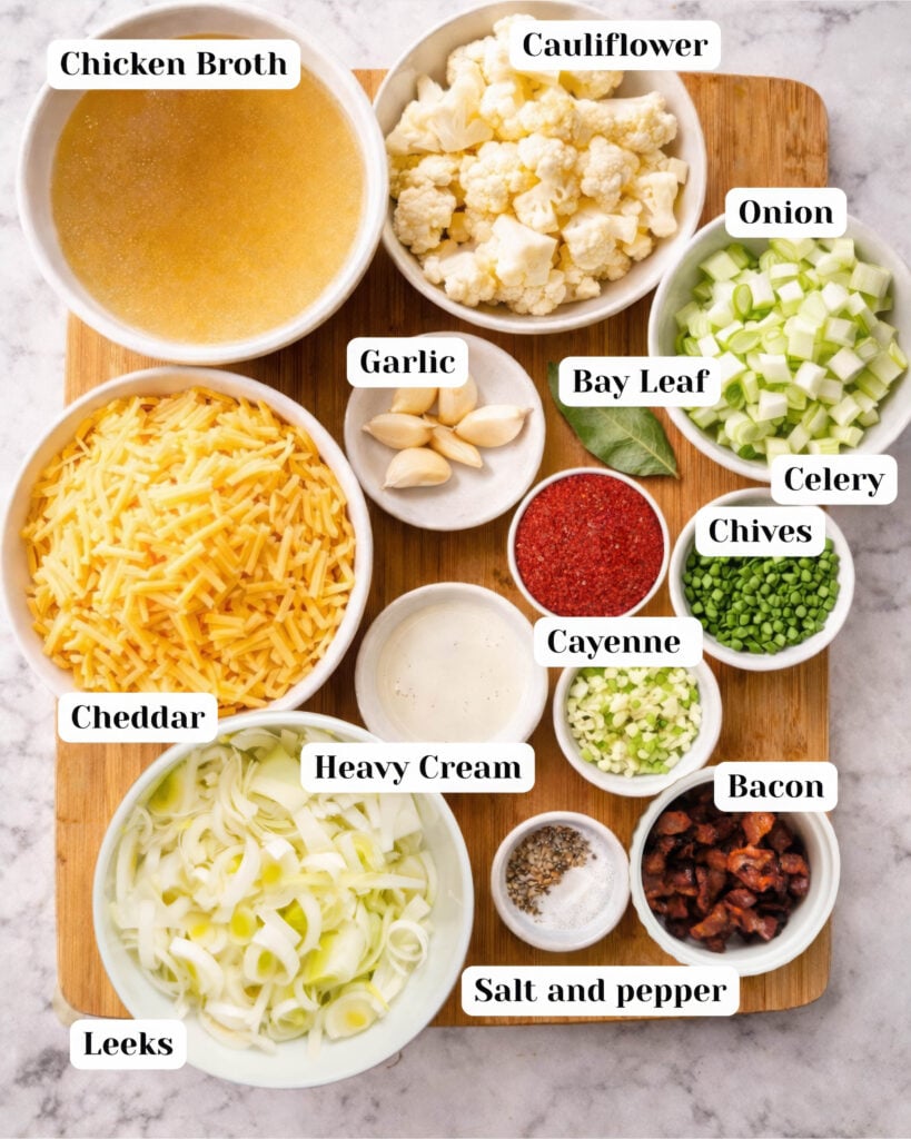 Overhead ingredient layout for crockpot cauliflower soup showing bowls of cauliflower florets, chicken broth, leeks, onion, celery, garlic, bay leaf, cheddar cheese, heavy cream, bacon, chives, cayenne, and salt and pepper on a wooden board.