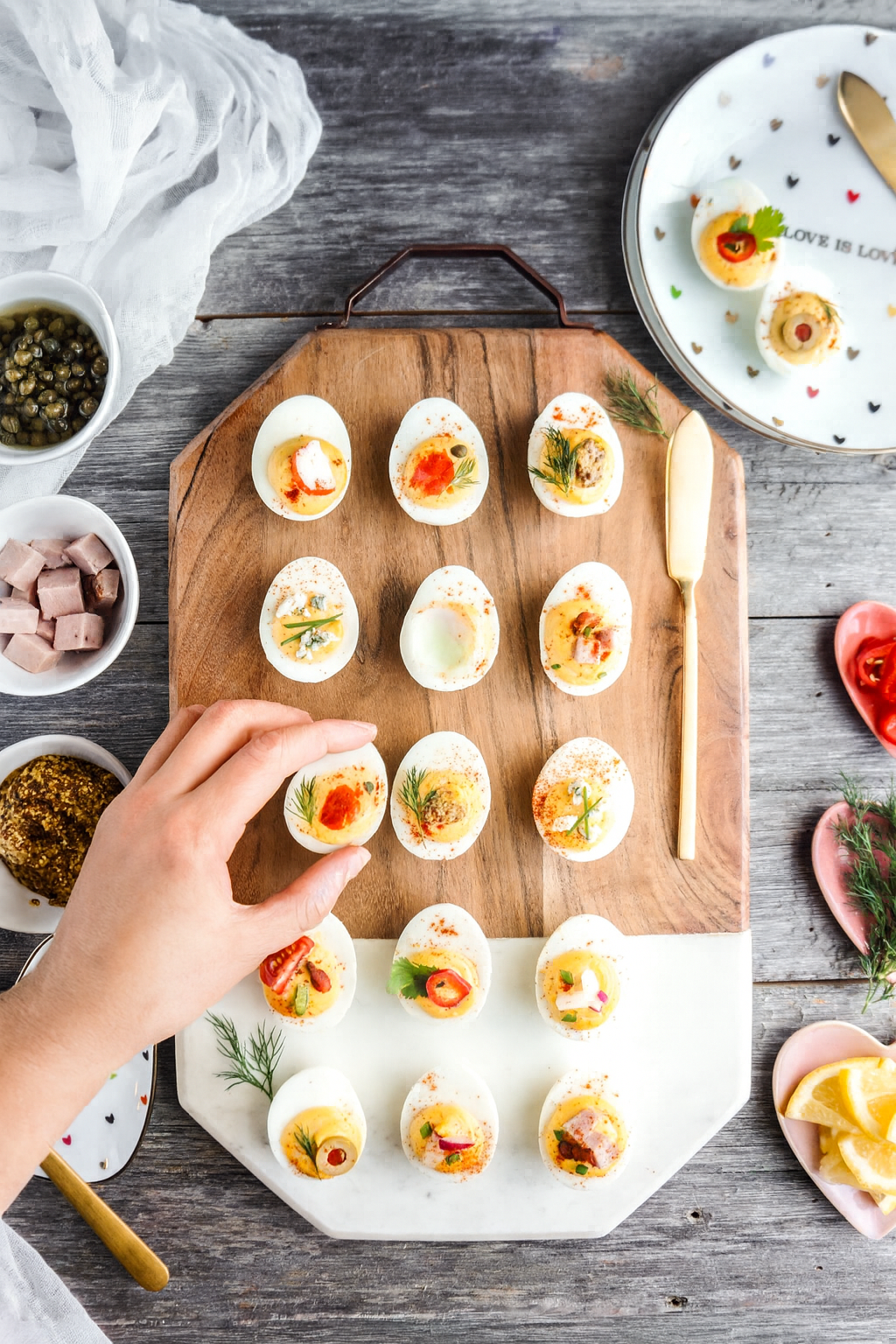 A top-down view of a colorful deviled egg platter arranged on a wooden and marble serving board, featuring a variety of garnished deviled eggs topped with herbs, peppers, bacon, and smoked salmon; a hand reaches in to grab one egg, surrounded by small bowls of capers, mustard, and ham on a rustic wooden table.
