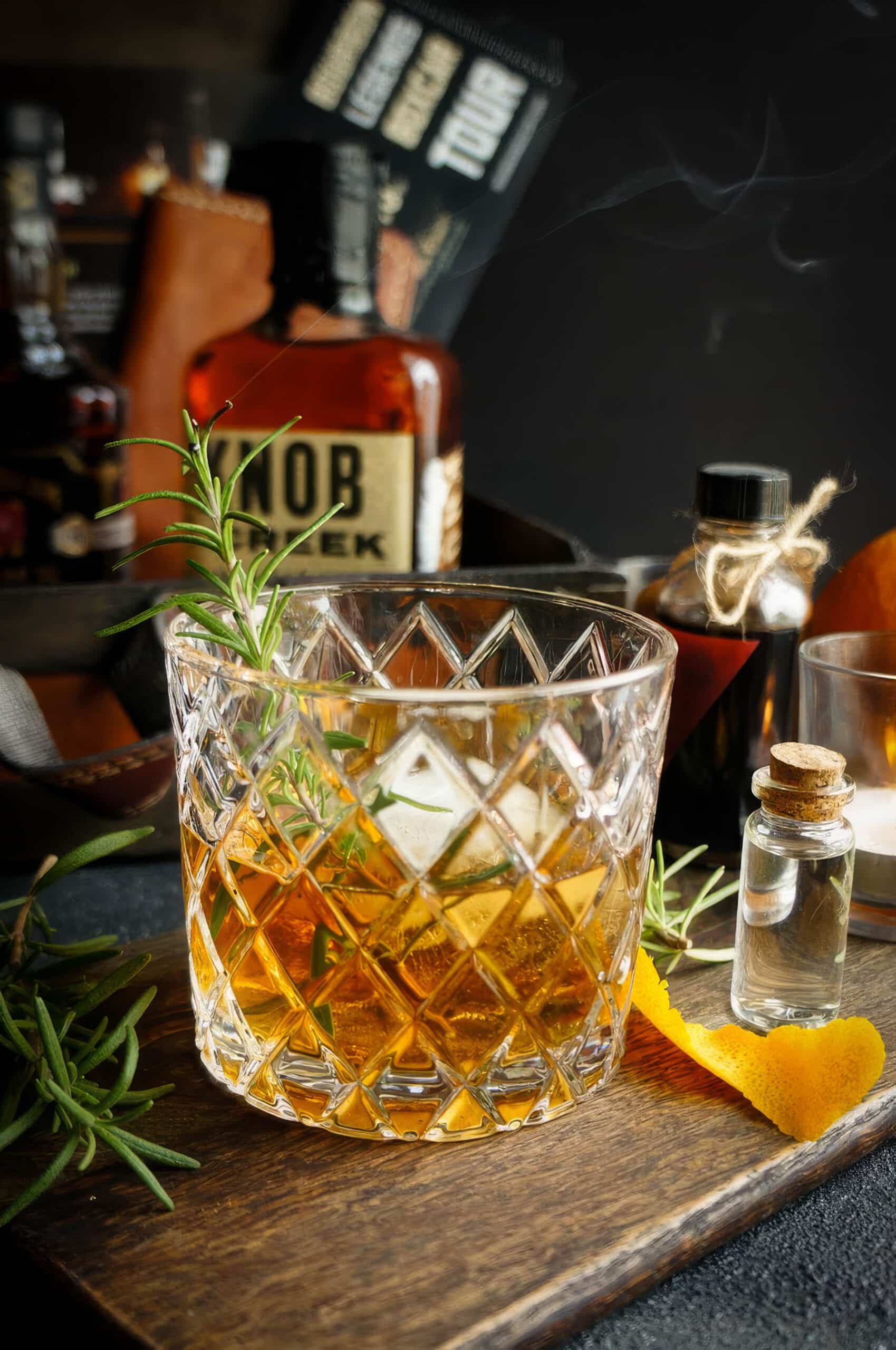 Rosemary bourbon old fashioned cocktail served over a large ice cube in a cut crystal rocks glass, garnished with a fresh rosemary sprig and orange peel on a dark wooden board.