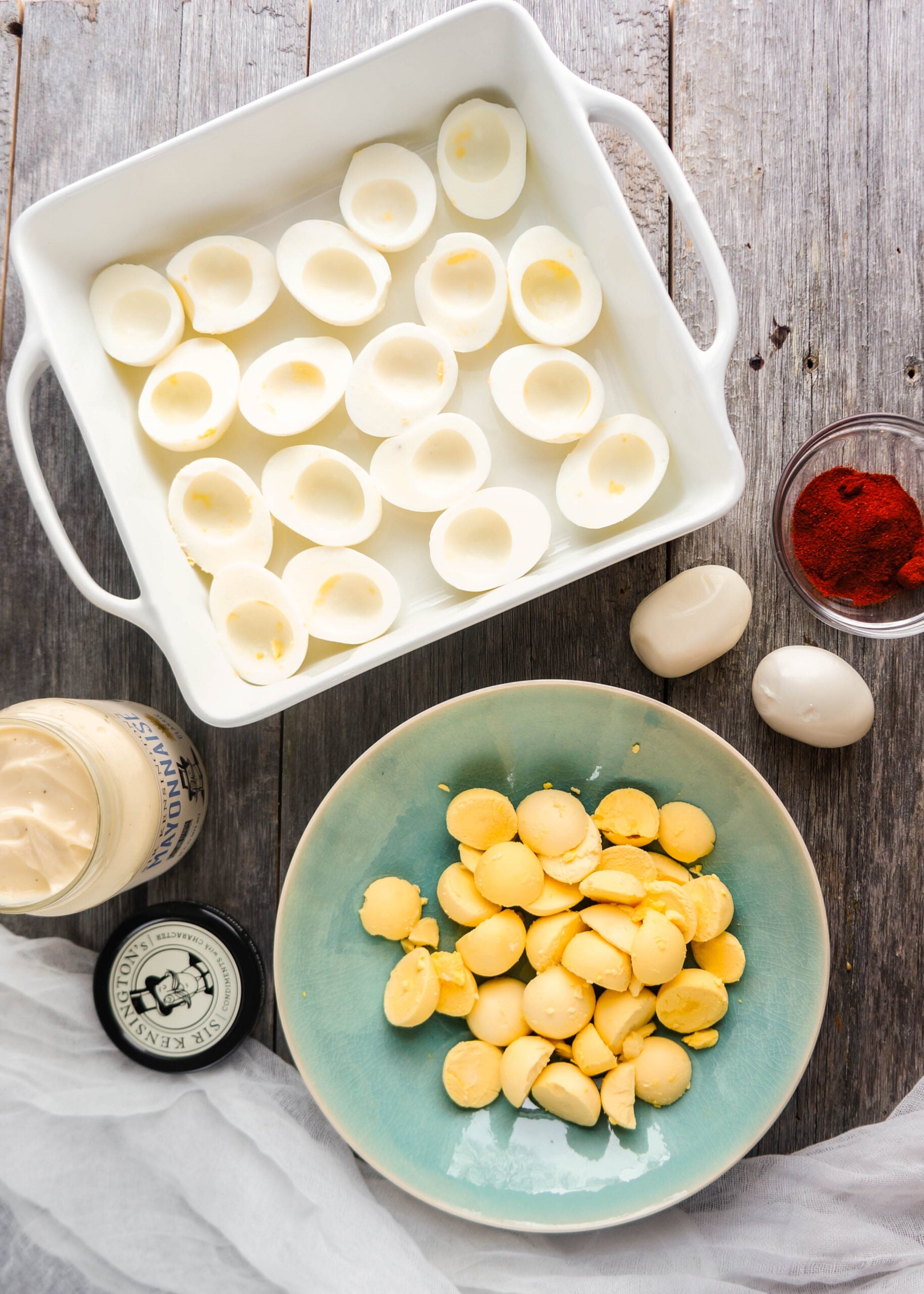Ingredients for a deviled egg platter arranged on a rustic wooden surface, including a white dish filled with halved hard-boiled egg whites, a turquoise bowl piled with bright yellow cooked yolks, a jar of mayonnaise with its lid beside it, a small bowl of paprika, and two whole peeled eggs, all set up for making deviled egg filling.