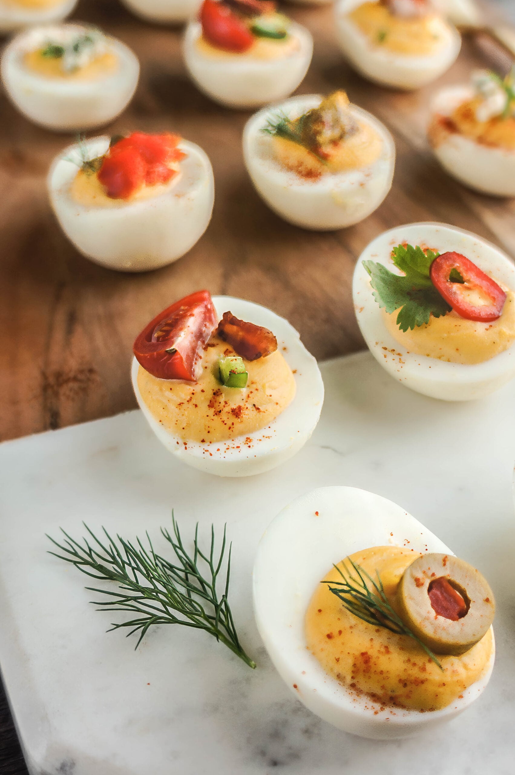 Close-up of a deviled egg platter featuring assorted toppings, including sliced tomatoes with bacon pieces, red chili peppers with cilantro, smoked salmon, blue cheese, dill, and stuffed olives, all arranged on a wooden board and marble slab for a colorful, elegant presentation.
