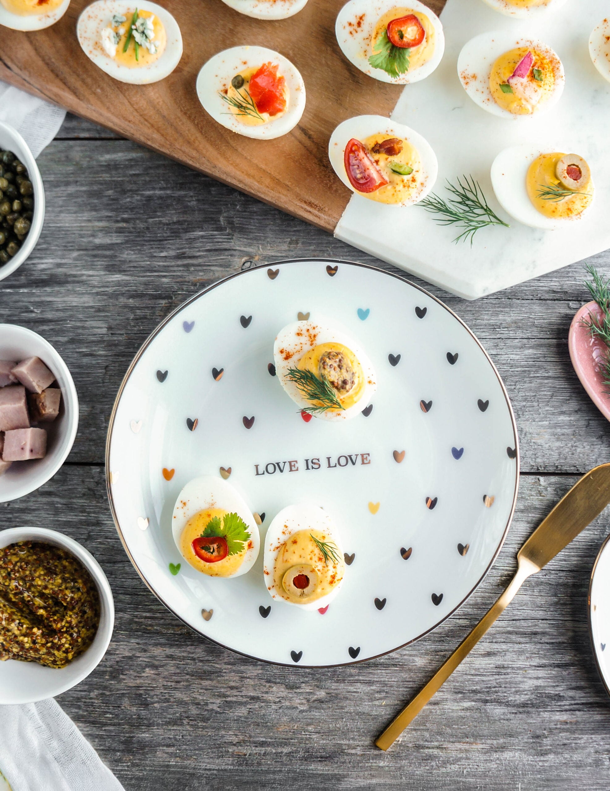 A colorful deviled egg platter displayed on a wooden and marble board, featuring assorted toppings like smoked salmon, dill, capers, blue cheese, bacon, tomatoes, chili peppers, and herbs. Three deviled eggs are plated on a white dish decorated with tiny multicolored hearts and the phrase ‘LOVE IS LOVE,’ surrounded by small bowls of diced ham, whole-grain mustard, and capers. A gold butter knife rests alongside the plate on a rustic wooden surface