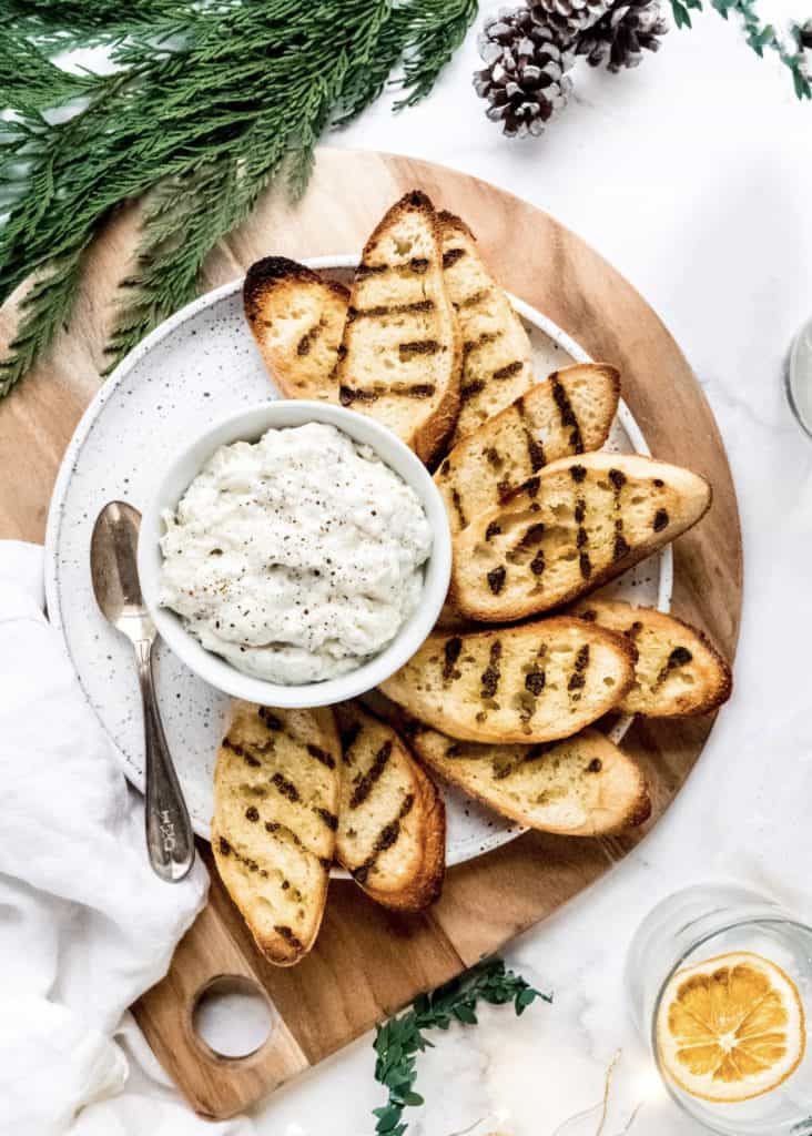 artichoke dip in bowl on plate with grilled baguette slices surrounding.