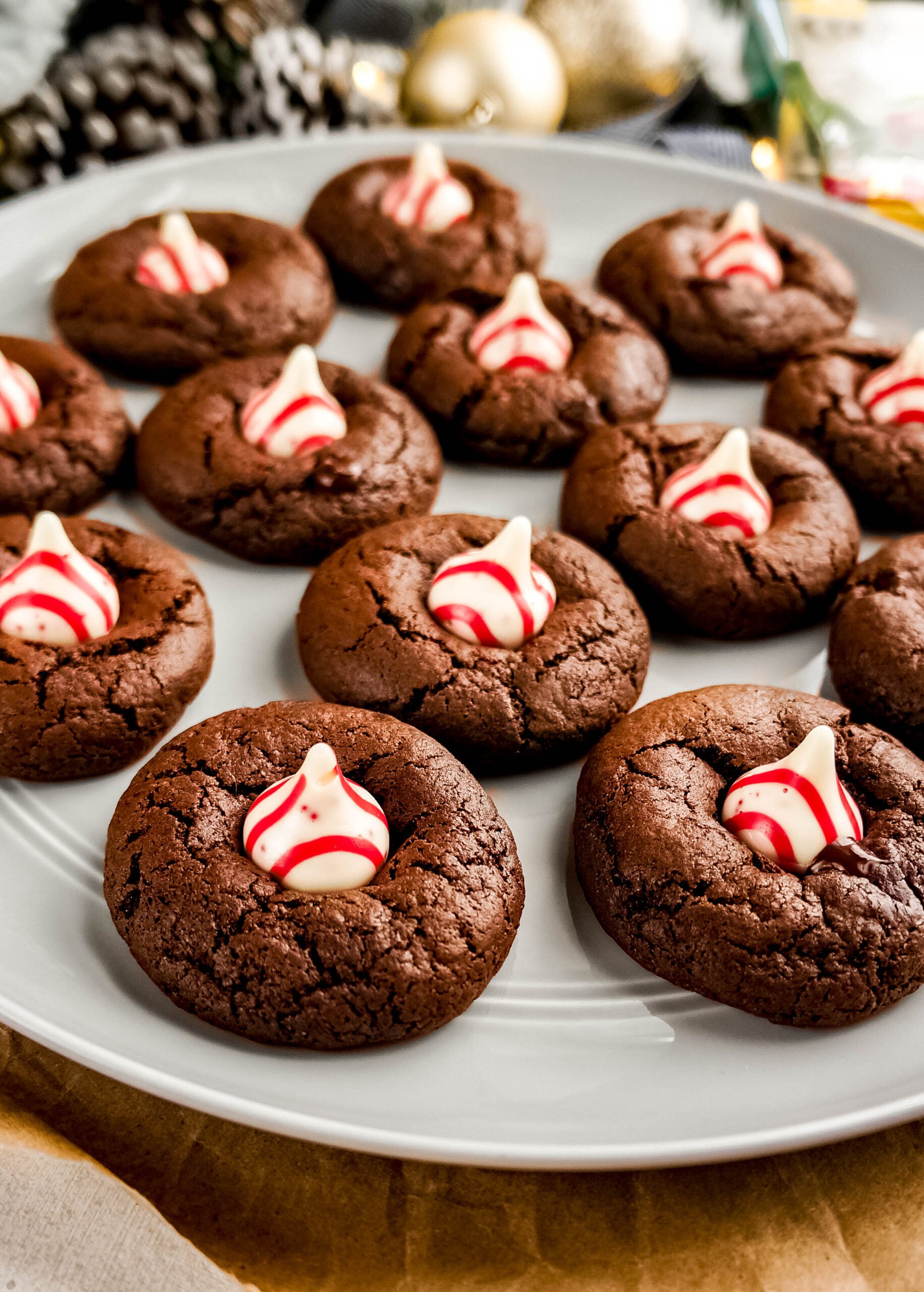 A white plate filled with soft, fudgy Peppermint chocolate kiss cookies, each topped with a red-and-white striped peppermint chocolate kiss. The rich chocolate cookies have crackled tops and the festive peppermint centers add a bright holiday pop, with Christmas décor blurred in the background.