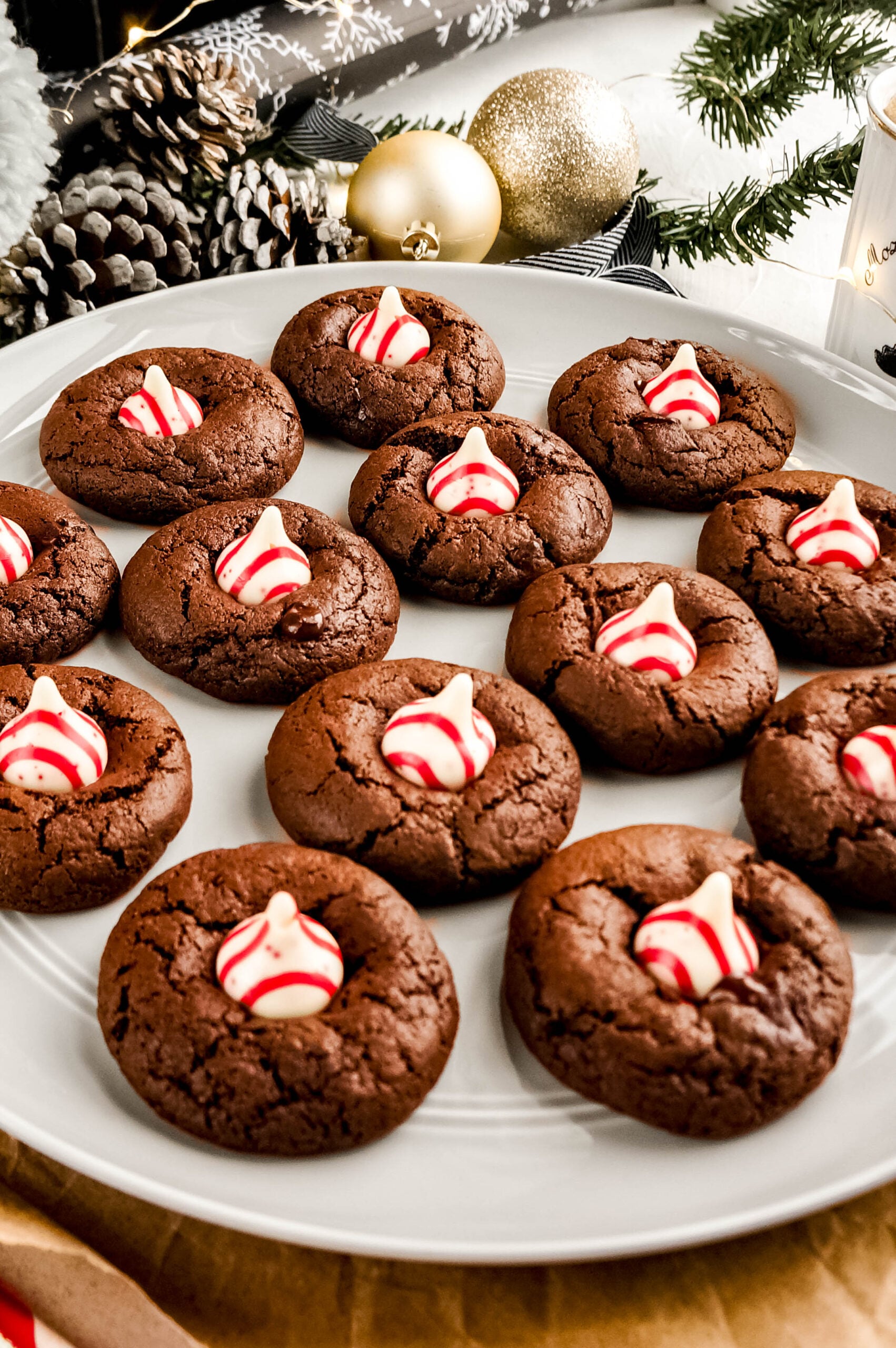 A white plate filled with soft, fudgy Peppermint chocolate kiss cookies, each topped with a red-and-white striped peppermint chocolate kiss. The rich chocolate cookies have crackled tops and the festive peppermint centers add a bright holiday pop, with Christmas décor blurred in the background.