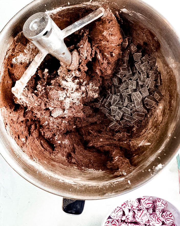 Chocolate cookie dough for Peppermint chocolate kiss cookies being mixed in a stand mixer bowl, with a pile of chocolate chunks ready to be stirred in. A small bowl of unwrapped peppermint chocolate kisses sits nearby, showing the ingredients in progress.