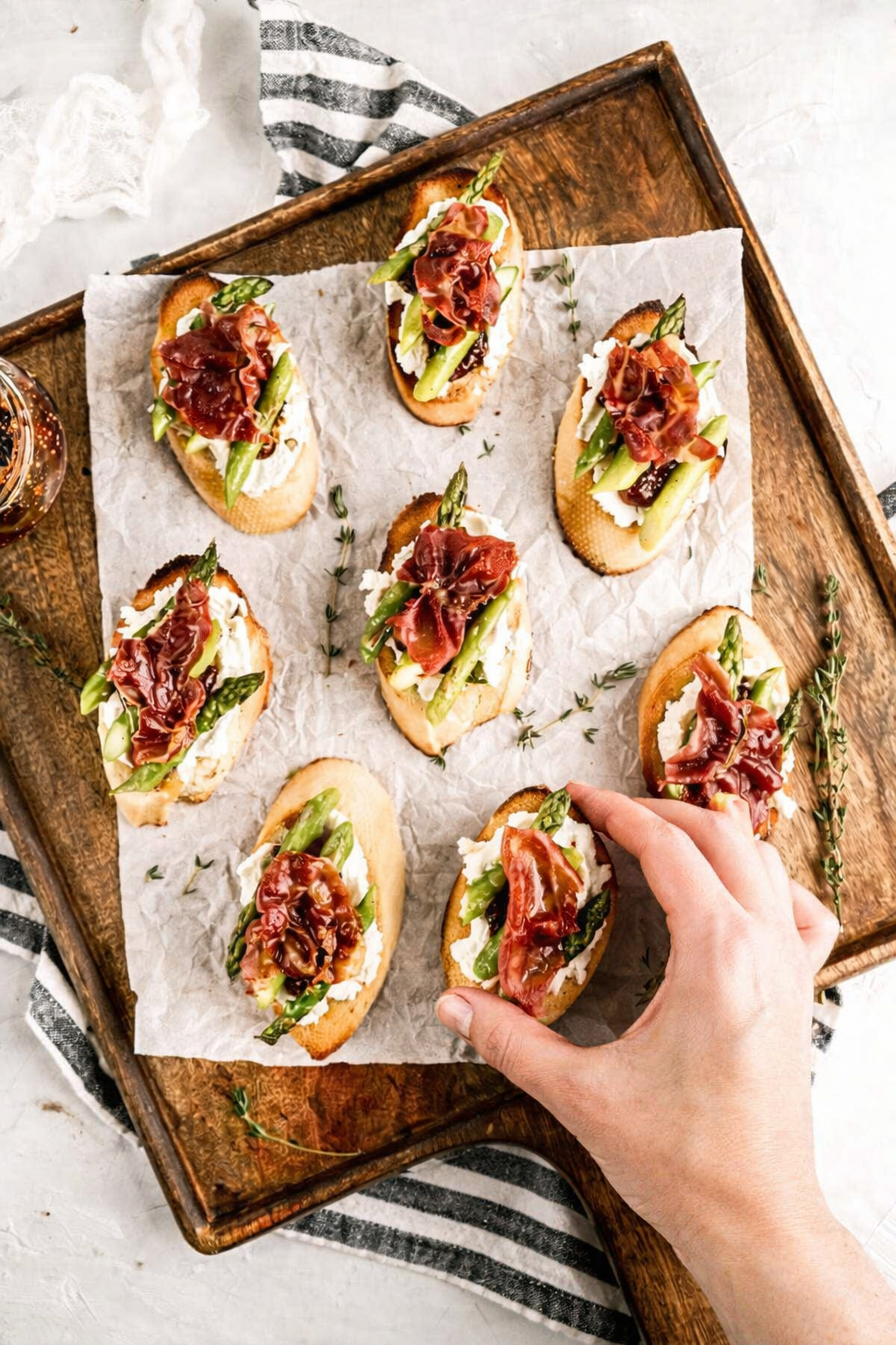 Overhead view of prosciutto crostini topped with creamy goat cheese, roasted asparagus, and fig jam on a wooden cutting board, with a hand reaching in to grab one crostini for serving.