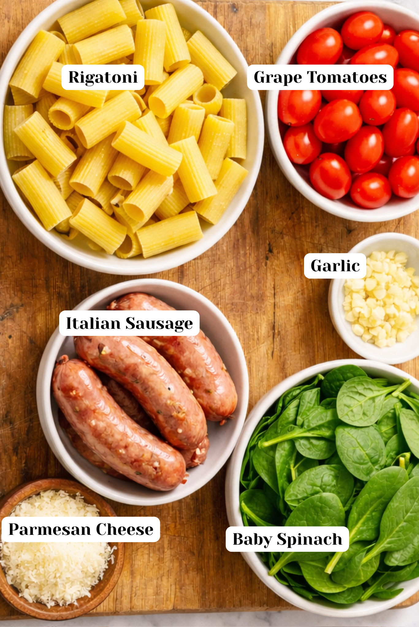 Quick and Easy Rigatoni with Sausage and Tomatoes Overhead view of ingredients for rigatoni with sausage and tomatoes, including rigatoni pasta, Italian sausage in casings, grape tomatoes, minced garlic, baby spinach, and freshly grated Parmesan cheese arranged in individual bowls on a wooden cutting board.