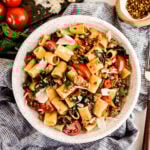 Overhead view of rigatoni with sausage and tomatoes in a white bowl, tossed with wilted spinach, juicy cherry tomatoes, and Italian sausage, finished with shaved Parmesan and red pepper flakes on a light marble surface.