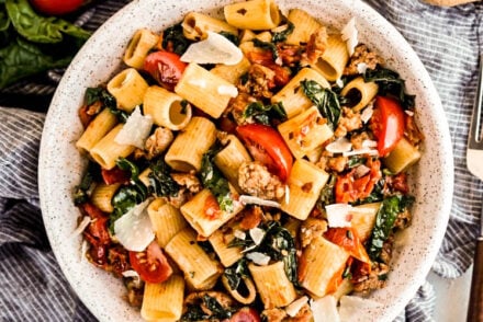 Overhead view of rigatoni with sausage and tomatoes in a white bowl, tossed with wilted spinach, juicy cherry tomatoes, and Italian sausage, finished with shaved Parmesan and red pepper flakes on a light marble surface.