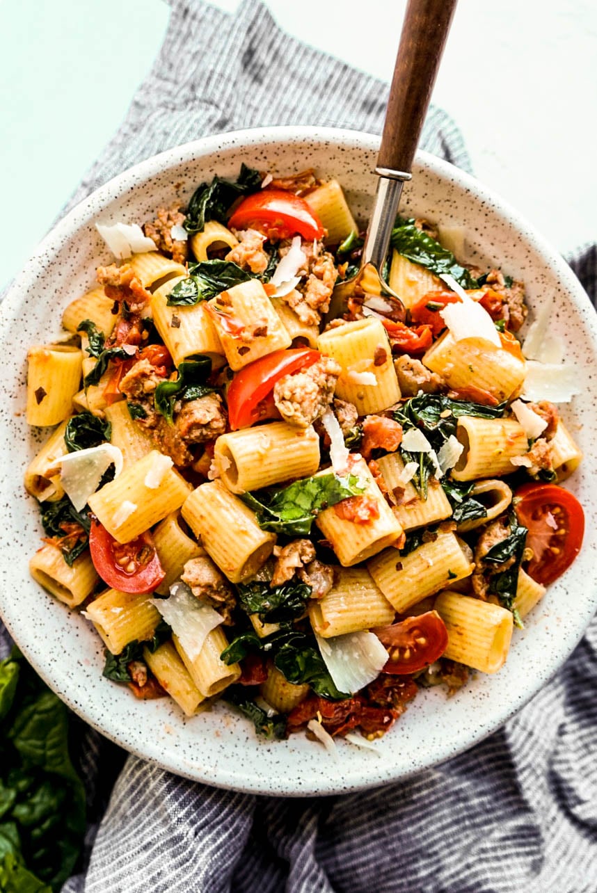 Overhead view of rigatoni with sausage and tomatoes, featuring tender pasta tossed with Italian sausage, burst cherry tomatoes, wilted spinach, and shaved Parmesan cheese.