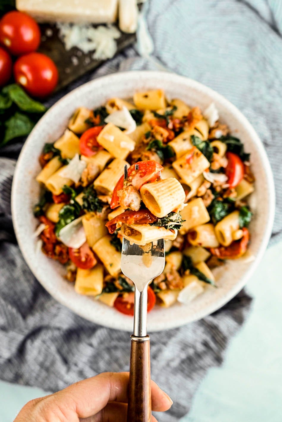 Fork lifting a bite of rigatoni with sausage and tomatoes, featuring tender pasta, Italian sausage, wilted spinach, and cherry tomatoes over a bowl of pasta.