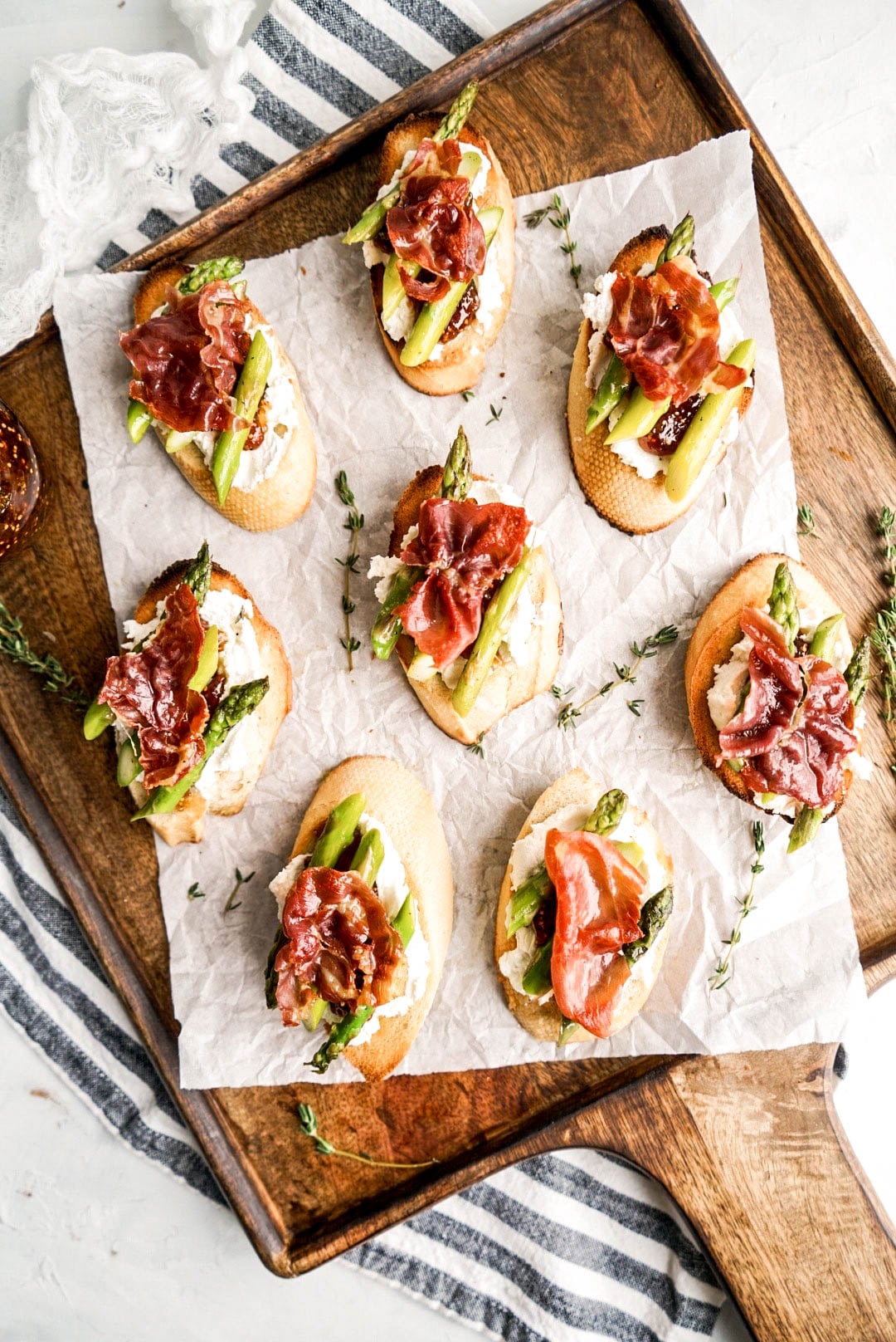 Overhead view of prosciutto crostini topped with creamy goat cheese, roasted asparagus, and fig jam, arranged on parchment paper over a wooden cutting board for an elegant appetizer spread.