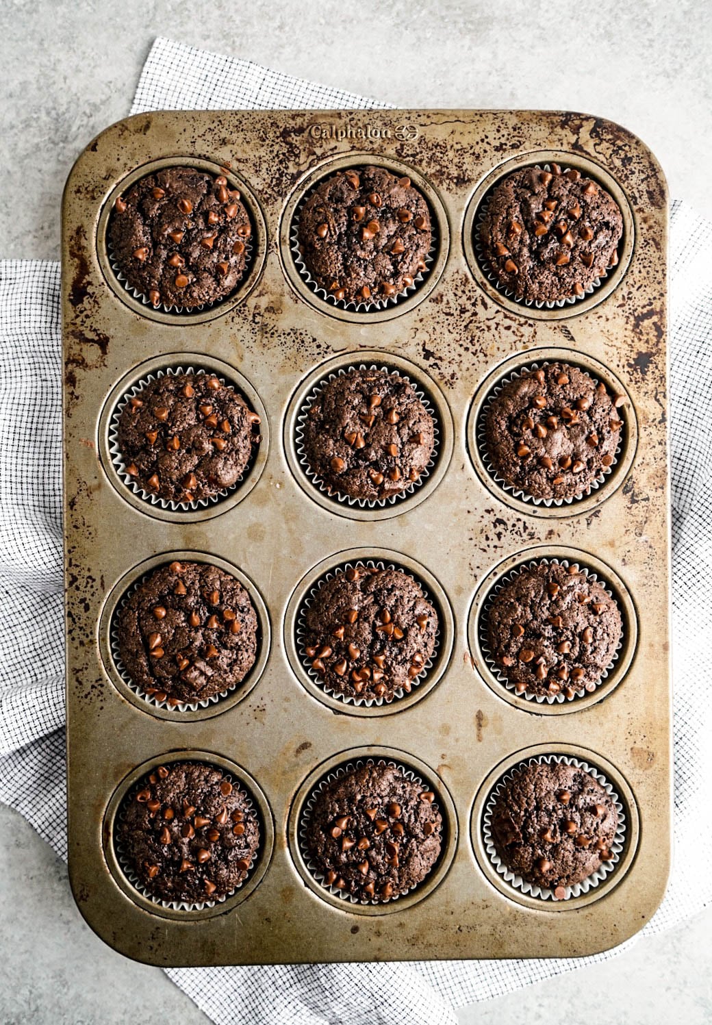 Overhead view of freshly baked Chocolate Zucchini Muffins studded with mini chocolate chips, baked in a rustic metal muffin tin and arranged on a light, textured surface with a linen towel.
