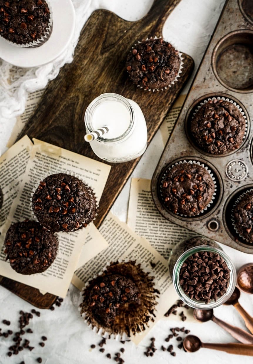 Overhead view of freshly baked Chocolate Zucchini Muffins topped with mini chocolate chips, styled in a rustic muffin tin and on a wooden cutting board with vintage book pages, scattered chocolate chips, copper measuring spoons, and a glass of milk on a light background.