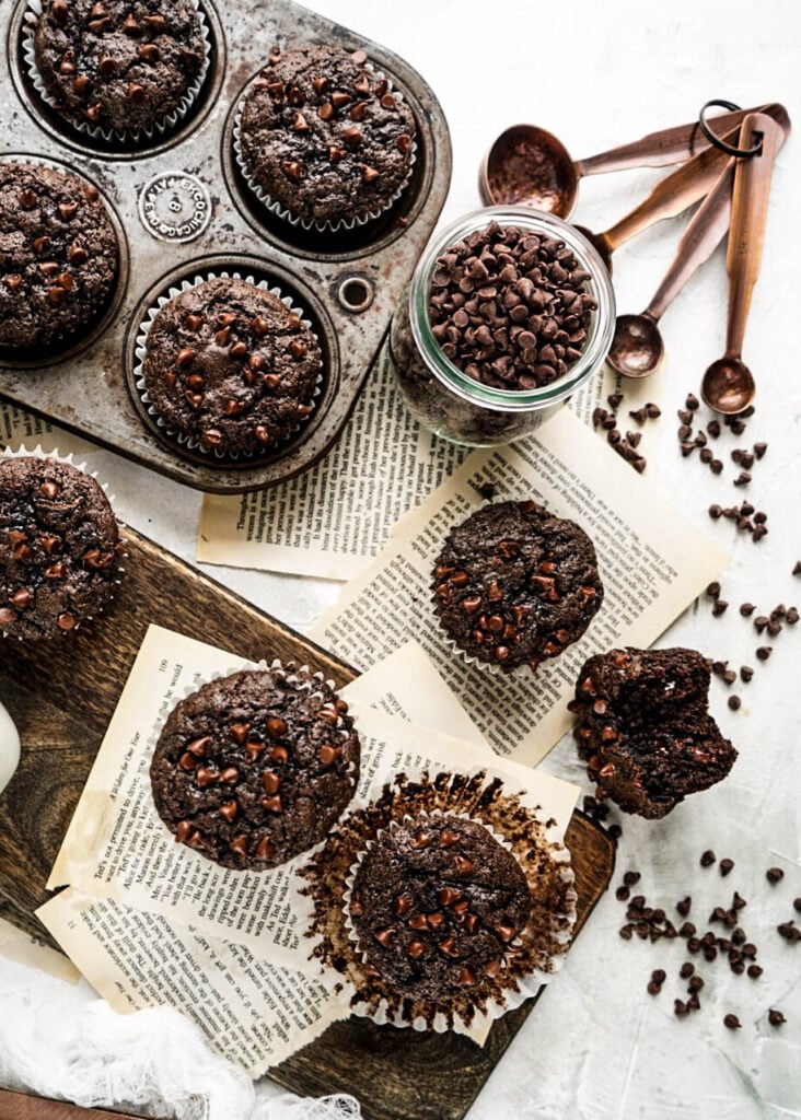 Overhead view of freshly baked Chocolate Zucchini Muffins topped with mini chocolate chips, arranged in a rustic muffin tin and on a wooden board with vintage book pages, scattered chocolate chips, and copper measuring spoons on a light background.
