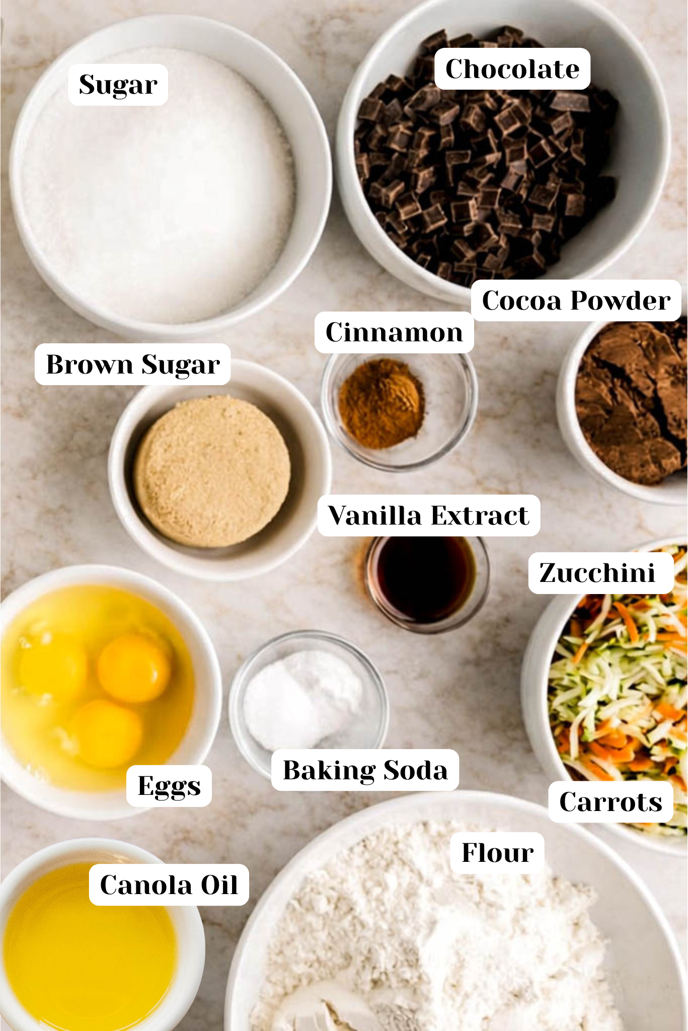 Overhead view of labeled ingredients for Chocolate Zucchini Muffins, including sugar, brown sugar, cocoa powder, chocolate chunks, cinnamon, vanilla extract, eggs, canola oil, baking soda, flour, grated zucchini, and shredded carrots arranged in white bowls on a light marble background.