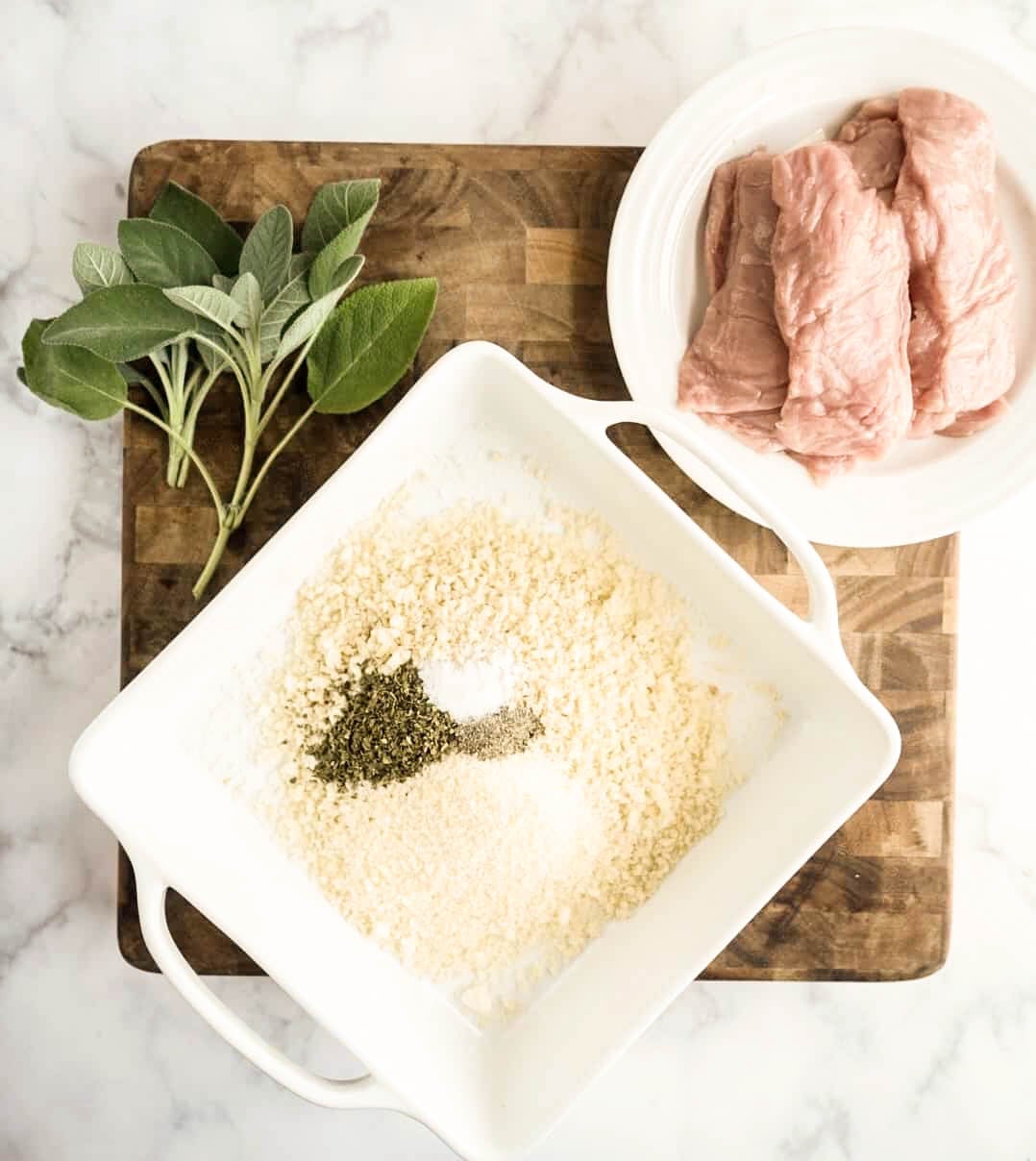 saltimbocca milanese Overhead view of panko breadcrumbs, Parmesan cheese, oregano, salt, and pepper in a white bowl with raw veal cutlets and fresh sage on a cutting board, ingredients prepared for making veal Milanese with Milanese sauce.