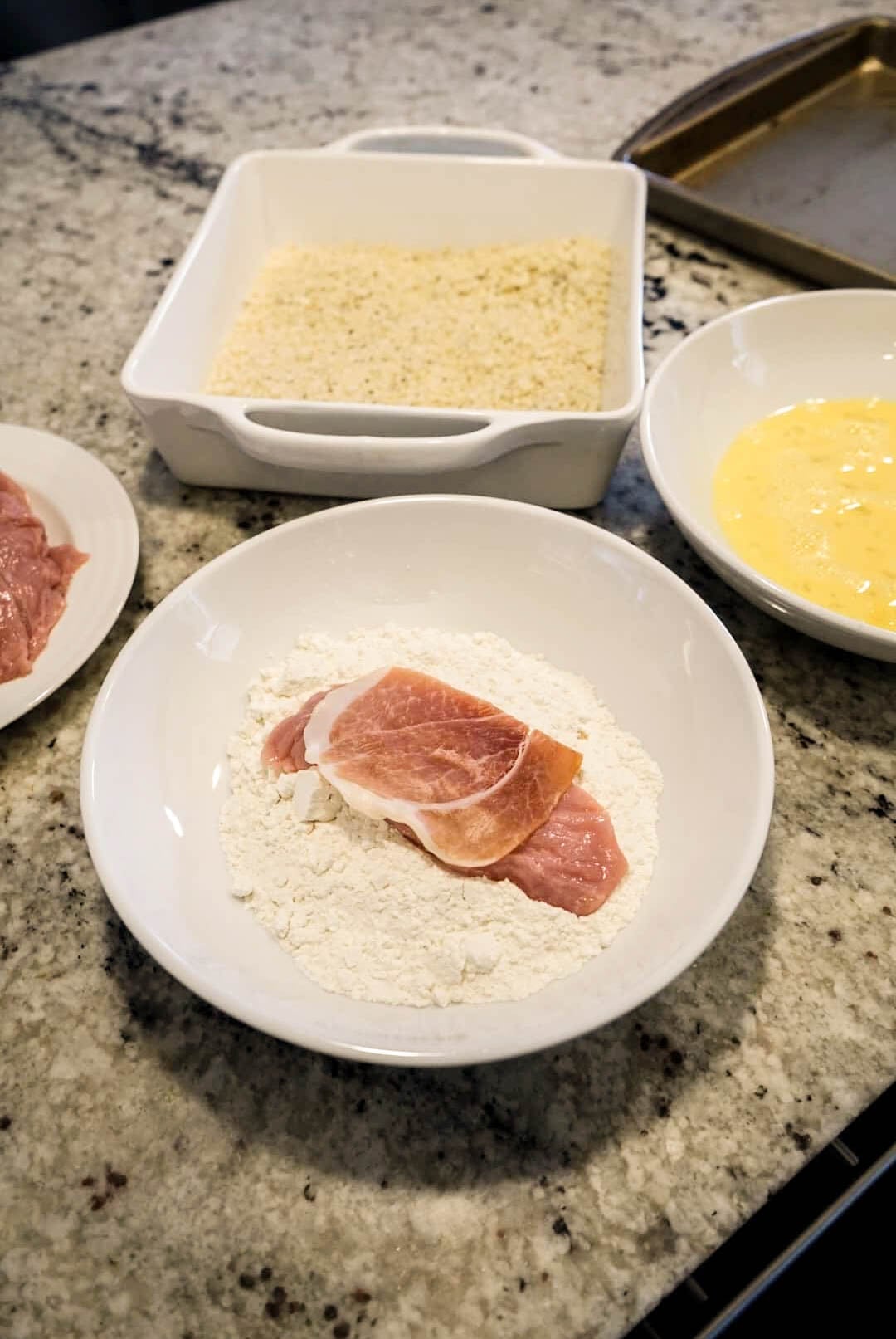 saltimbocca milanese Veal cutlet dredged in flour with prosciutto on top beside bowls of egg wash and panko breadcrumbs, part of the breading process for veal Milanese served with Milanese sauce.