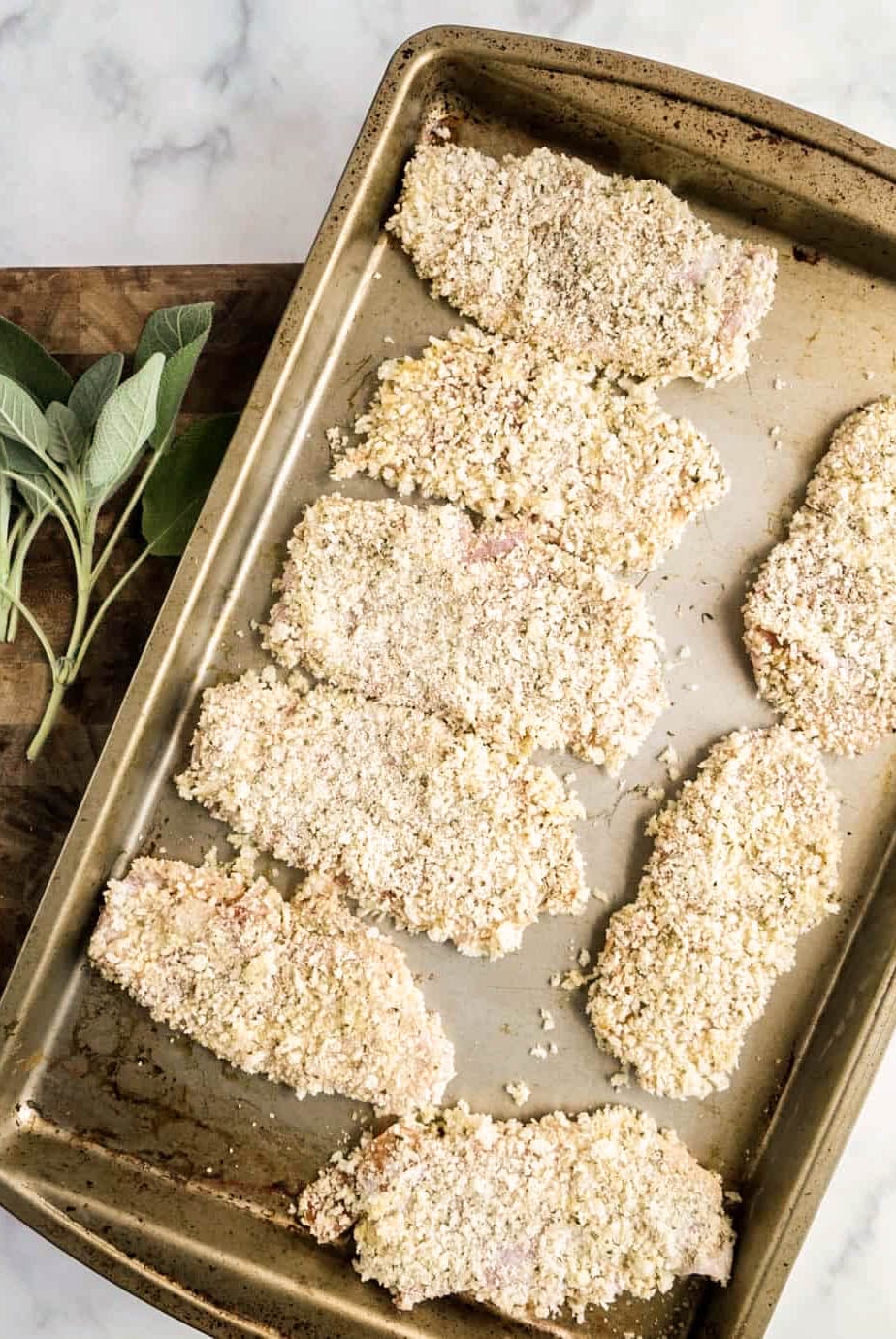 saltimbocca milanese Raw veal cutlets coated in panko and Parmesan breadcrumbs arranged on a sheet pan beside fresh sage leaves, prepared for frying and serving with Milanese sauce.