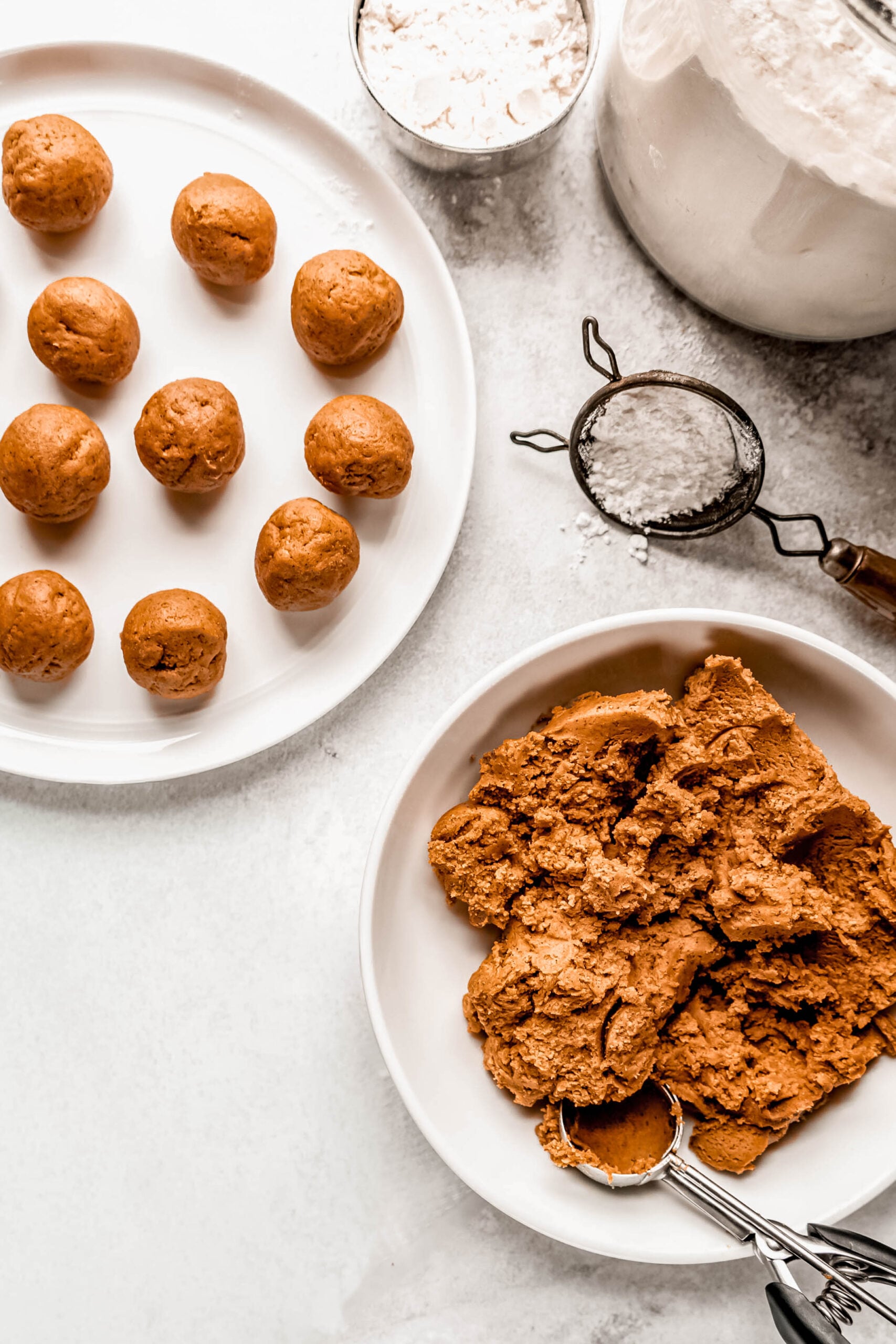 Balls of pfeffernusse cookie dough rest on a white plate beside a large bowl filled with spiced dough and a cookie scoop. A sifter and a container of flour sit nearby on a light surface, showing the preparation process before baking