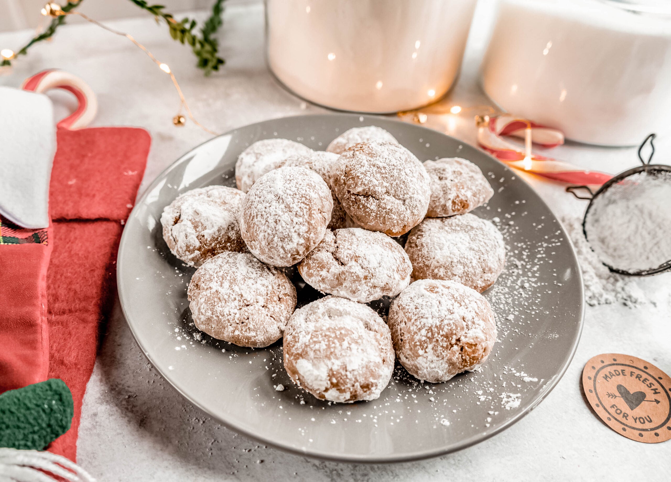 A plate piled high with powdered sugar–coated pfeffernüsse cookies sits on a holiday-themed table, surrounded by festive décor like candy canes, twinkle lights, and a sifter dusted with sugar.