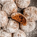A close-up of powdered sugar–coated pfeffernüsse cookies on a plate, with one cookie broken open to show its soft, spiced interior.