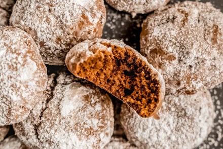 A close-up of powdered sugar–coated pfeffernüsse cookies on a plate, with one cookie broken open to show its soft, spiced interior.