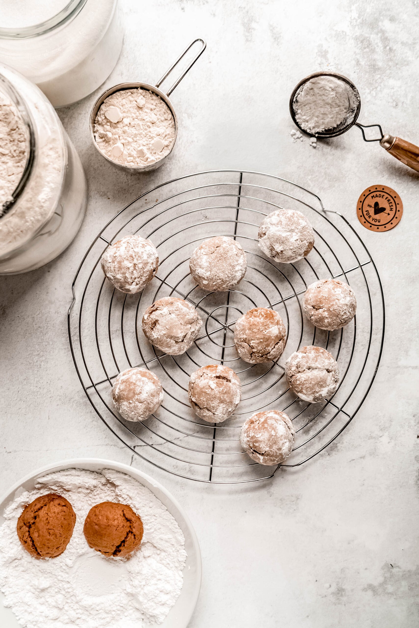 Freshly baked pfeffernüsse cookies coated in powdered sugar cool on a round wire rack, with extra dough balls resting in powdered sugar nearby. Measuring cups, flour jars, and a small sifter surround the setup, showing the cookie-coating process