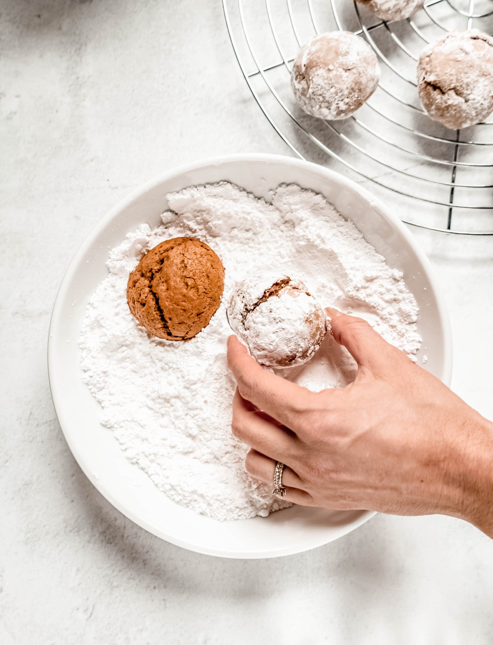 A hand rolls warm Pfeffernüsse cookies in a shallow bowl of powdered sugar, coating the spiced cookies while freshly dusted cookies cool on a wire rack in the background.