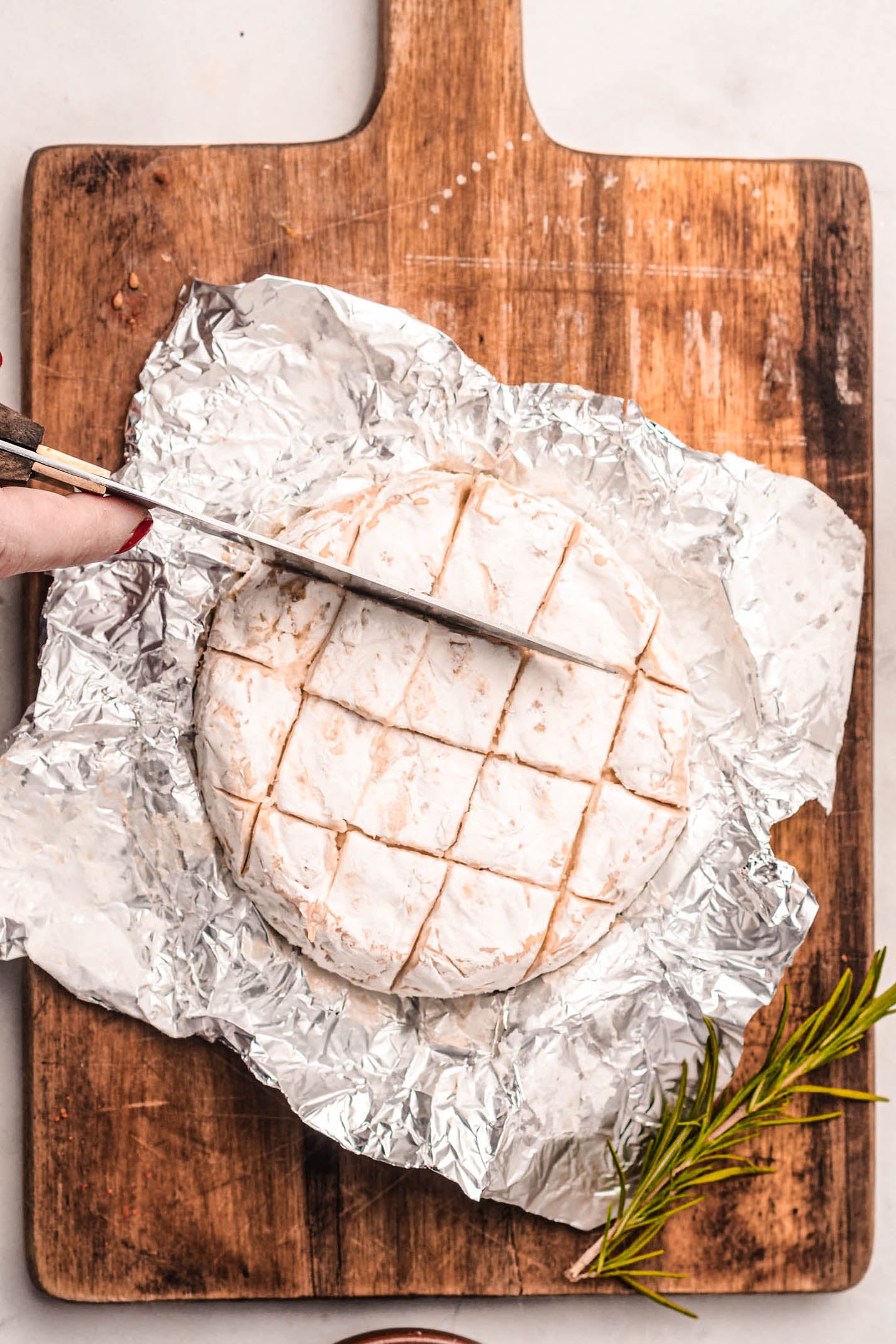 A round wheel of brie sits on a sheet of crinkled aluminum foil atop a rustic wooden cutting board. The brie rind is scored in a crosshatch pattern as a hand with red-polished nails slices through the top with a sharp knife. A fresh sprig of rosemary rests beside the cheese, hinting at the savory baked brie with caramelized onions it will become.