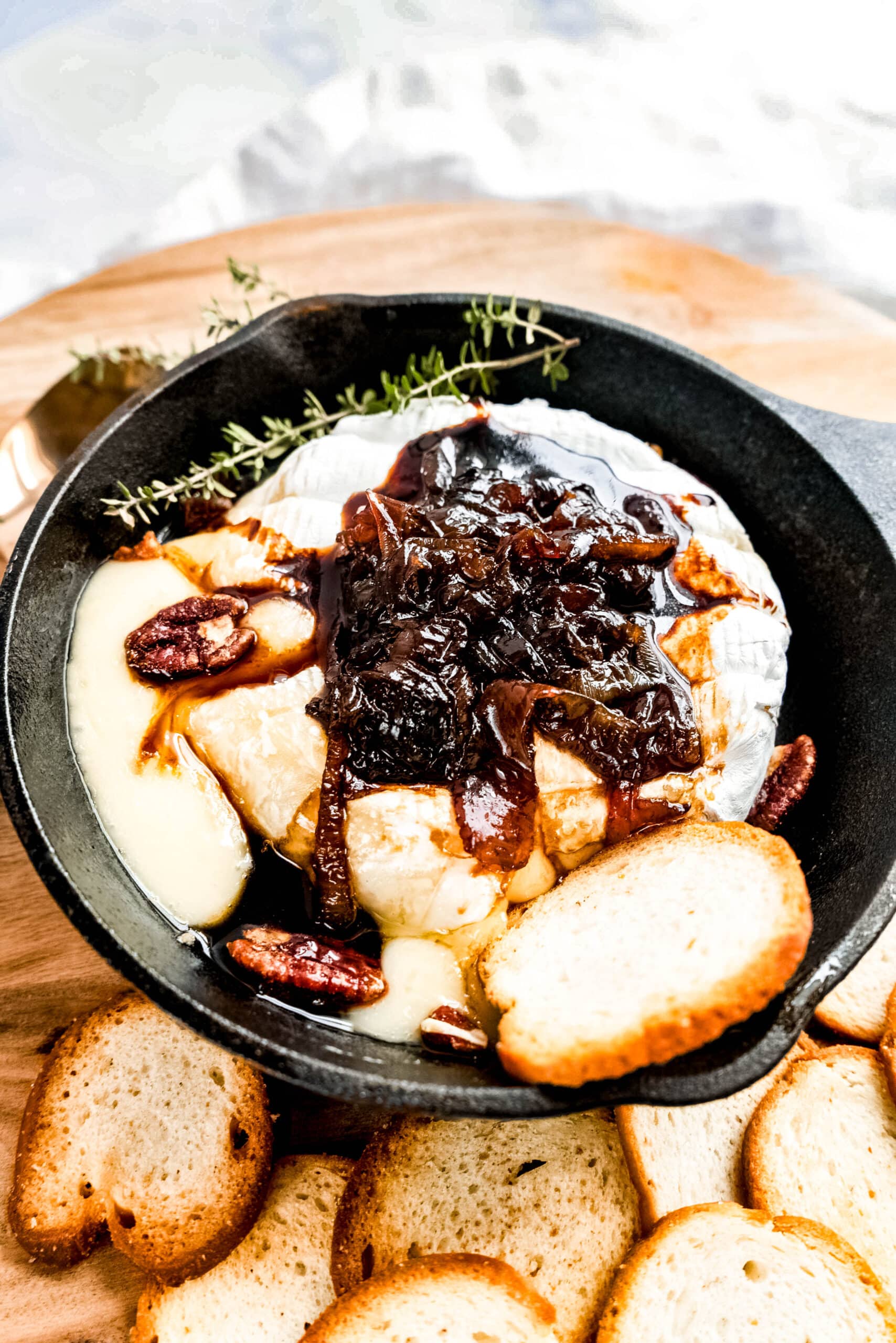 Warm Baked Brie with Caramelized Onions served in a cast-iron skillet, topped with glossy balsamic caramelized onions, fresh herbs, toasted pecans, and surrounded by crisp baguette slices for dipping.