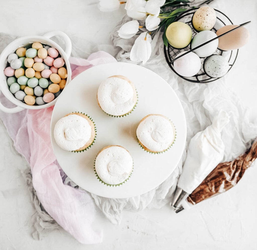 Easter egg cupcakes four baked easter egg cupcakes with frosting on a white cake stand with a bowl of candy eggs to the left, eggs in a basket to the right with white tulips and two bags of frosting on a white background