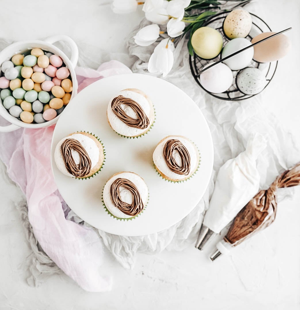 Easter egg cupcakes four baked easter egg cupcakes without frosting and nests on a white cake stand with a bowl of candy eggs to the left, eggs in a basket to the right with white tulips and two bags of frosting on a white background