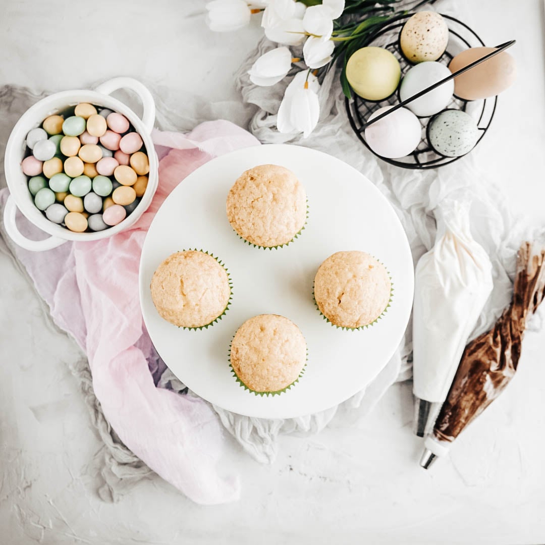 Easter egg cupcakes four baked easter egg cupcakes without frosting on a white cake stand with a bowl of candy eggs to the left, eggs in a basket to the right with white tulips and two bags of frosting on a white background