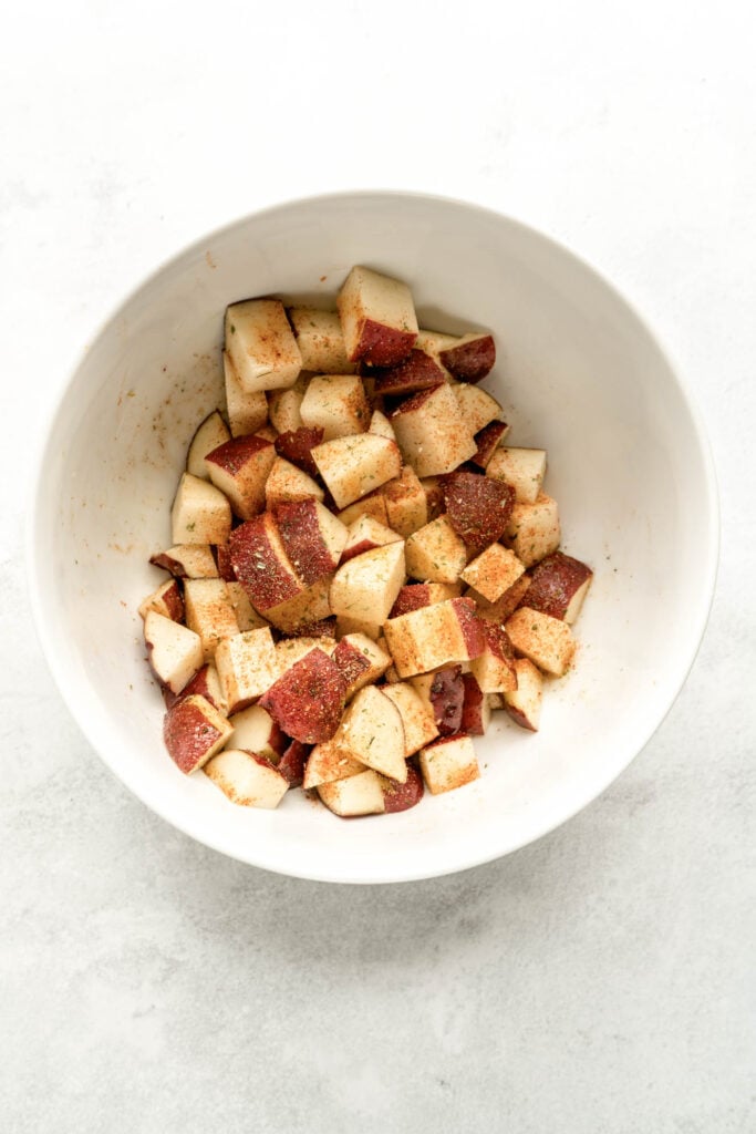 Cubed red potatoes tossed with olive oil and seasonings in a white mixing bowl, ready to be cooked into crispy air fryer cubed potatoes.
