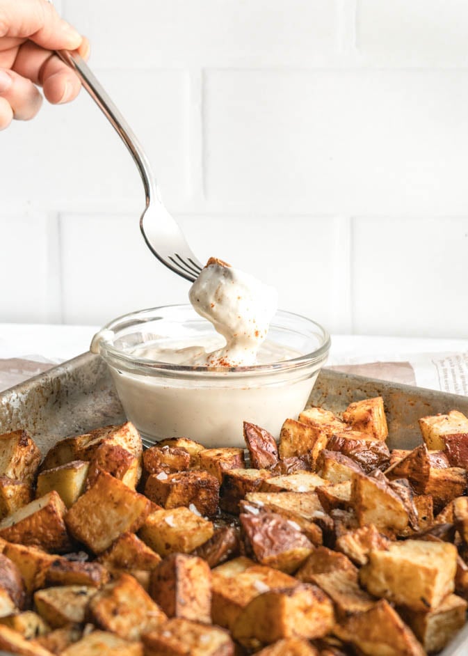 A hand holding a fork dips a crispy golden air fryer cubed potato into a small glass bowl of creamy truffle aioli sauce, surrounded by roasted potato cubes on a metal baking sheet.