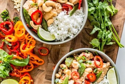 **Alt text:** *Overhead view of two bowls of healthy cashew chicken with white rice, cashews, red bell peppers, sugar snap peas, pineapple, and fresh cilantro on a wooden cutting board.*