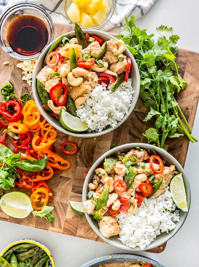 **Alt text:** *Overhead view of two bowls of healthy cashew chicken with white rice, cashews, red bell peppers, sugar snap peas, pineapple, and fresh cilantro on a wooden cutting board.*