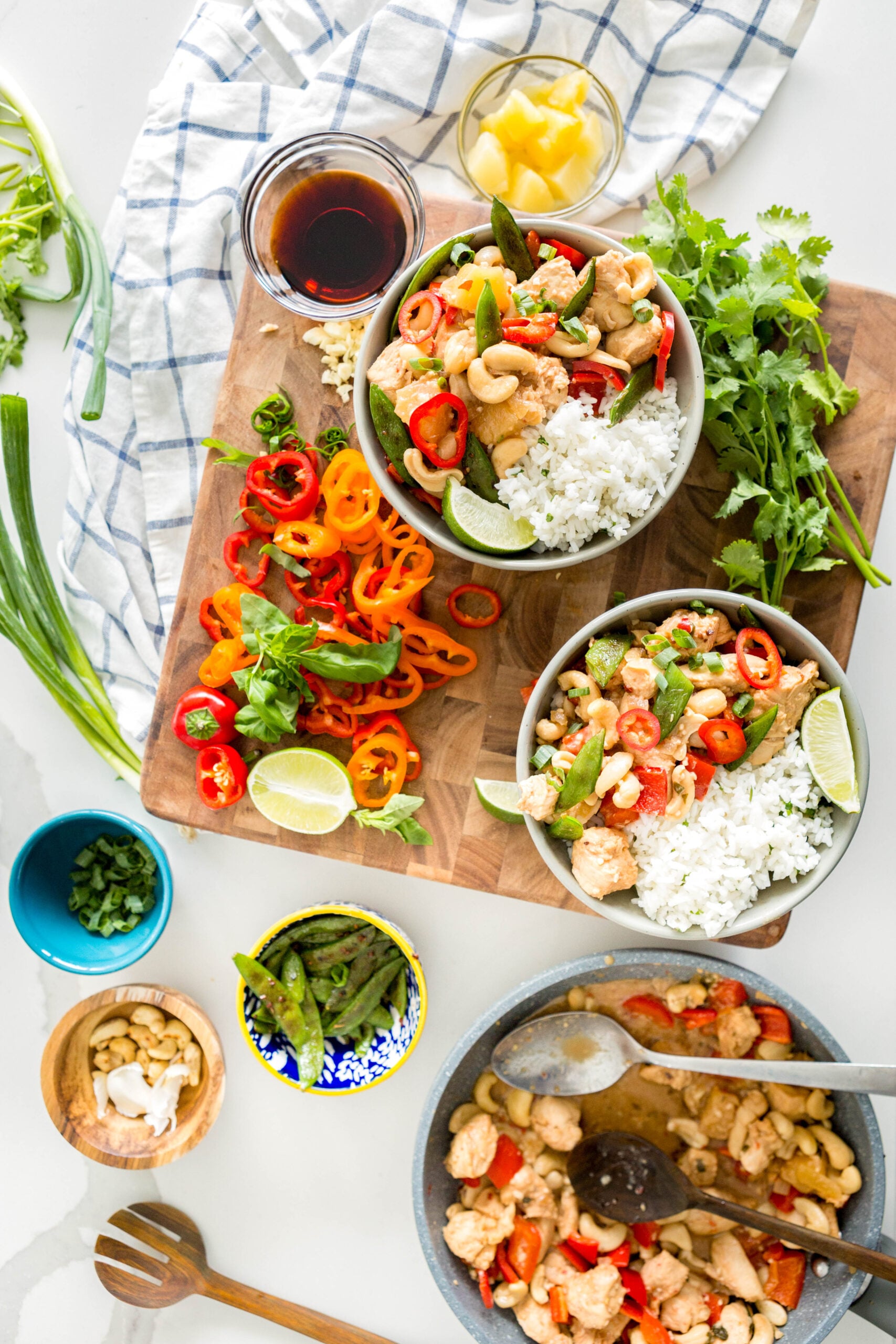**Alt text:**
Overhead view of two bowls of healthy cashew chicken with white rice, cashews, red bell peppers, sugar snap peas, pineapple, and fresh cilantro on a wooden cutting board.