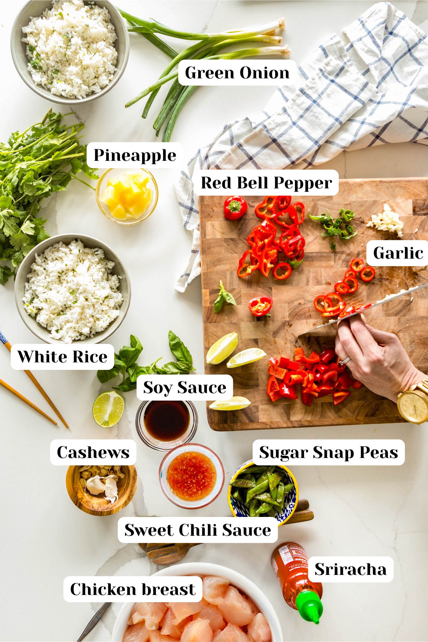 Overhead ingredient layout for healthy cashew chicken featuring chicken breast, white rice, pineapple, red bell pepper, sugar snap peas, cashews, garlic, green onion, soy sauce, sweet chili sauce, and sriracha on a white marble surface.
